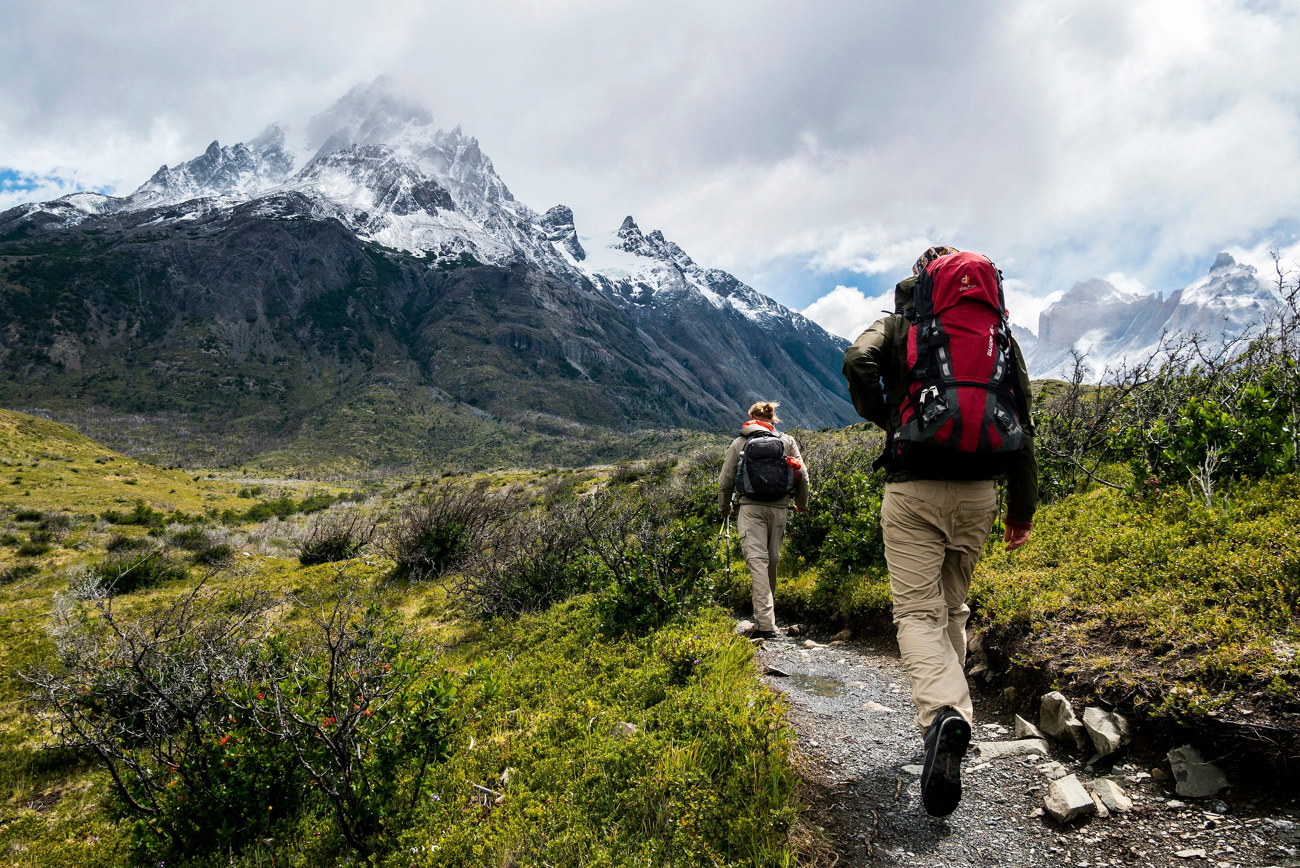 Two hikers trek across open terrain with large backpacks toward snow-capped mountain peaks.