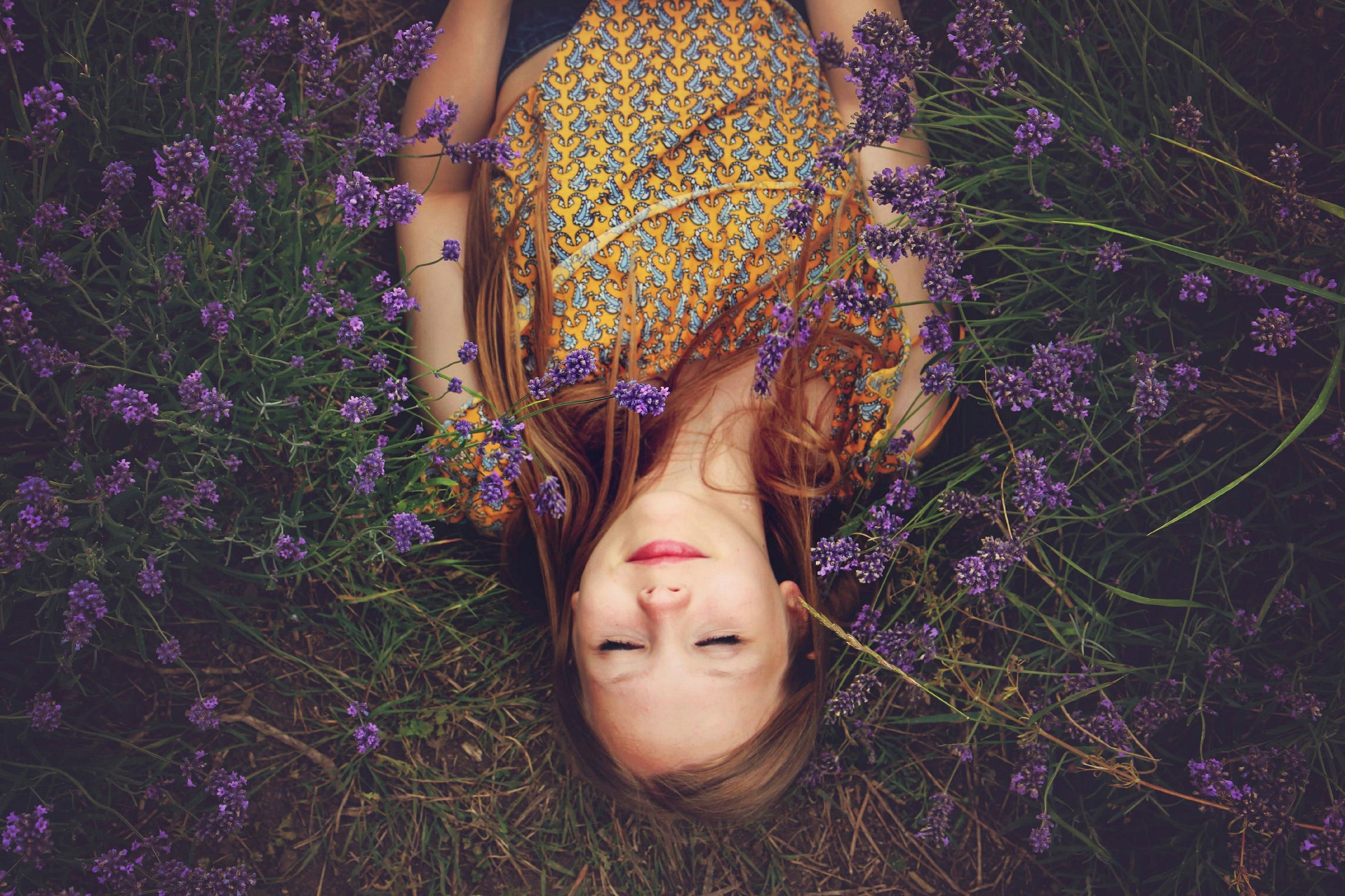 Girl in yellow patterned dress lying peacefully among purple lavender flowers with eyes closed.