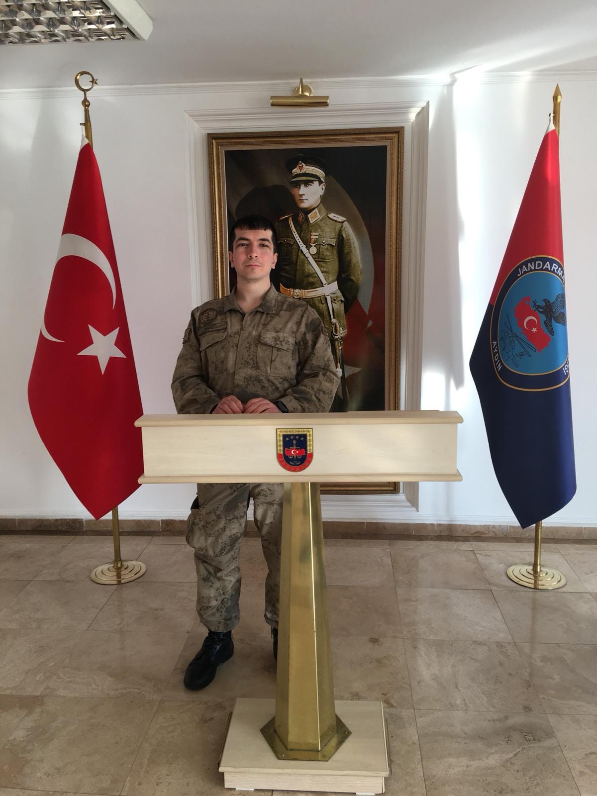 Young man in Turkish military uniform standing at a podium between Turkish flag and Gendarmerie flag in an official building, looking at camera.