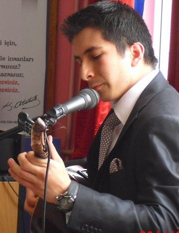 A young man speaking into a microphone with a red curtain and writing board visible in the background.