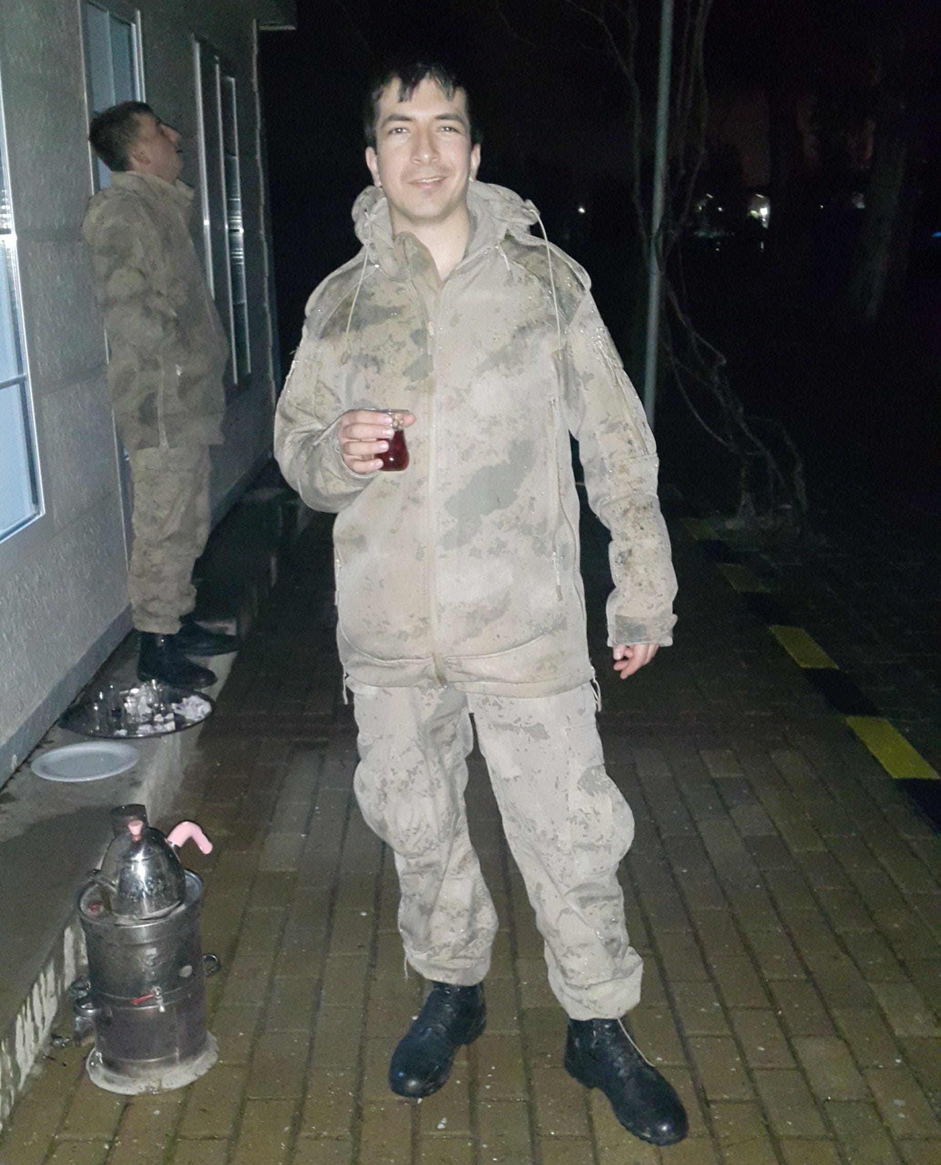 A young soldier in military uniform stands in a corridor holding a red beverage in his hand.