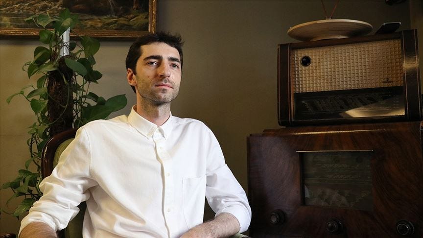 Beyaz gömlek giymiş, koyu saçlı bir adam vintage radyo ve bitki dekoru bulunan retro bir ortamda oturmuş, yanda bakıyor.