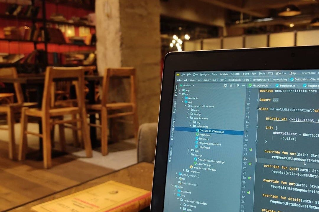 Android IDE'de Kotlin kodu ve proje dosya ağacı görüntüleyen geliştirici ekranı, arka planda bir kafe ortamı.