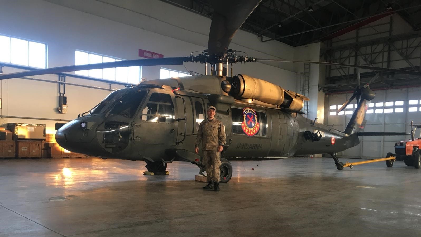 A soldier in military uniform stands beside a green Sikorsky Black Hawk helicopter parked in a hangar.
