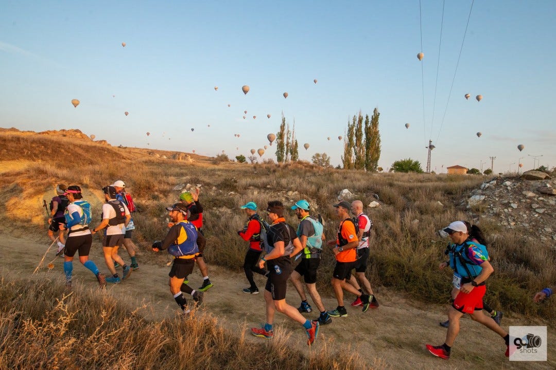 A group of runners exercising on hilly terrain with dozens of colorful hot air balloons flying in the sky above them.