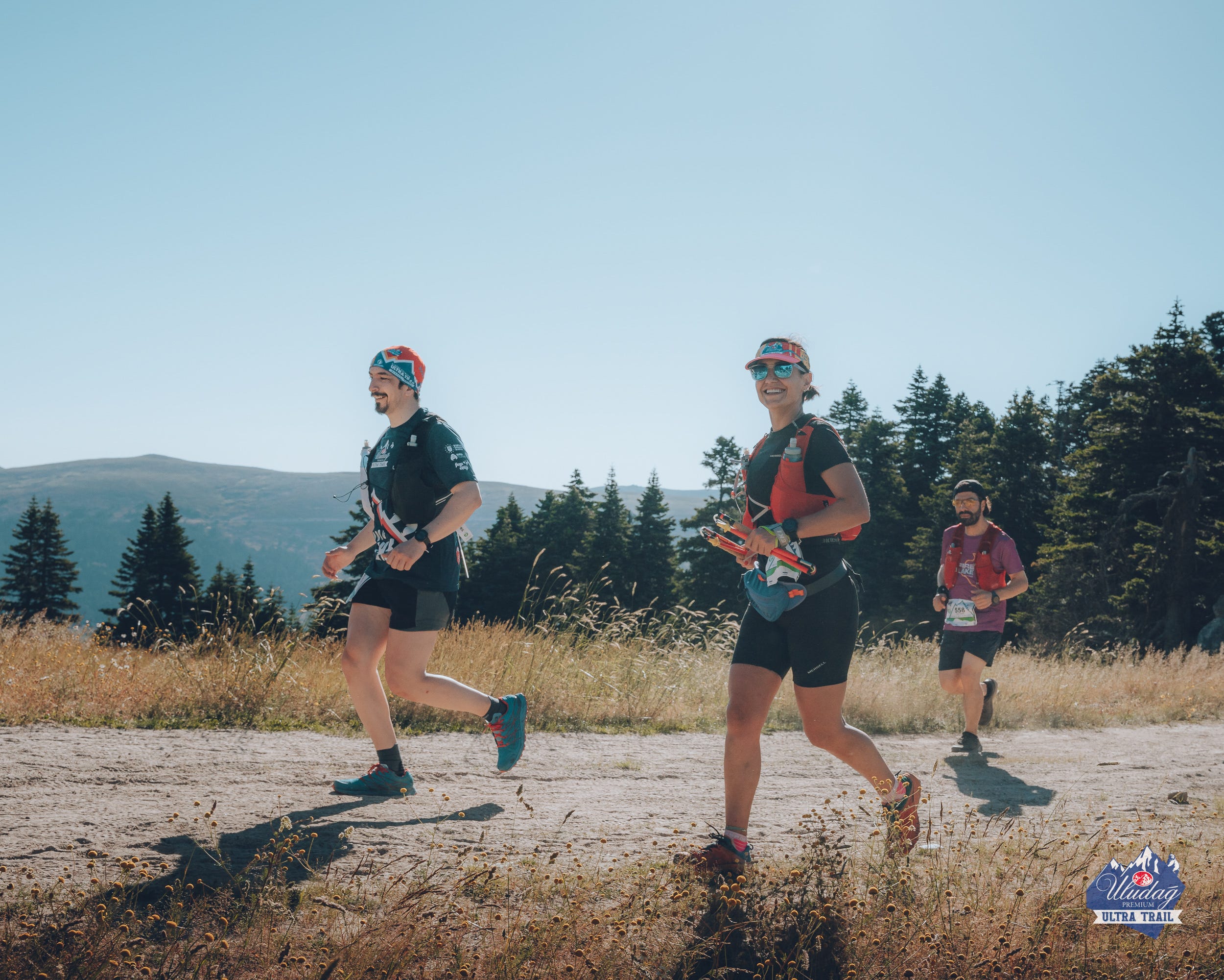 Three trail runners jogging across mountainous terrain under clear blue sky with forested hills in the background.