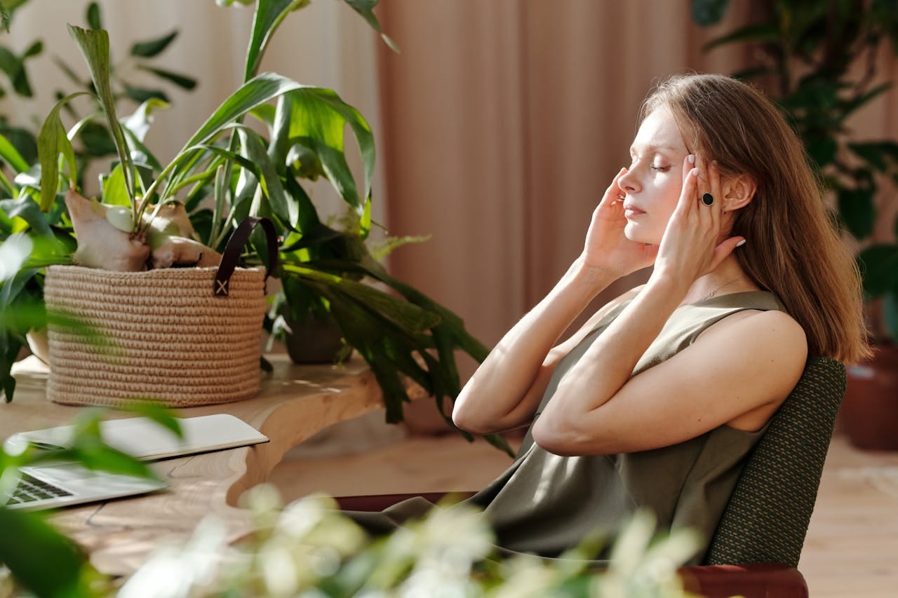 A woman at her desk with plants touches her face in pain, suffering from a headache.